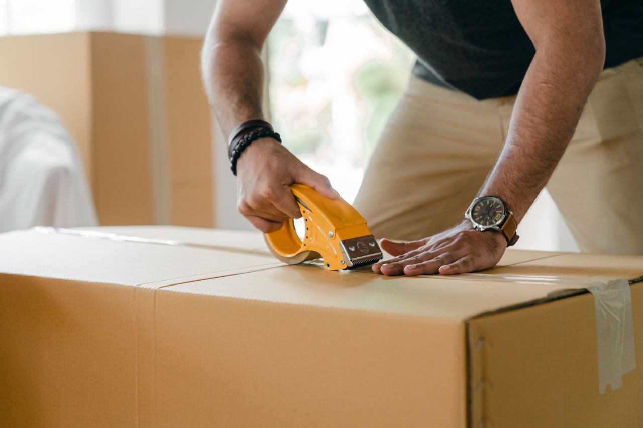 Crop unrecognizable young male in casual clothes packing cardboard box and sealing with scotch tape